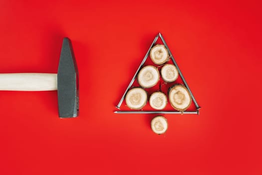 Long nails and wood cuts or branch slabs, laid out in shape of Christmas tree with hammer on red background. Flat lay