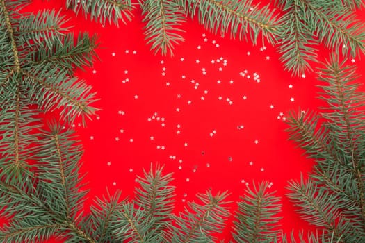Branches of a Christmas tree on a red background with silver confetti stars. Merry christmas and happy new year concept. Flat lay. Copy space.