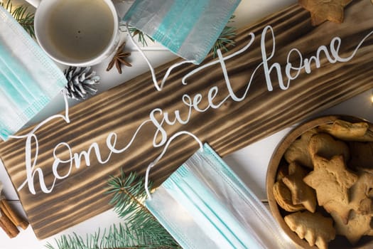 Home Sweet Home lettering burnt wood sign, gingerbread cookie, Christmas tree branches, cup of coffee, cinnamon sticks, medical masks on white surface. Flat lay. Quarantine during a pandemic concept