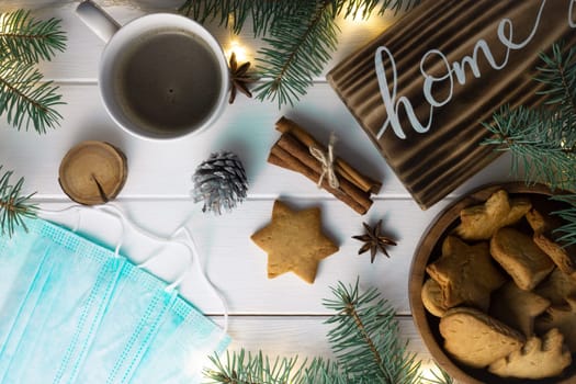 Home lettering wood sign with gingerbread cookie, Christmas tree branches, cup of coffee, cinnamon sticks and medical masks on white surface. Flat lay. Quarantine during a pandemic concept