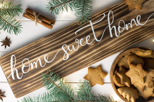 Home Sweet Home lettering burnt wood sign with gingerbread cookie, Christmas tree branches, cinnamon sticks and anise on white surface. Flat lay