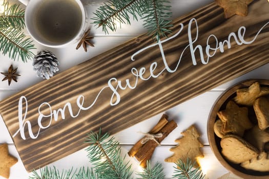 Home Sweet Home lettering burnt wood sign, gingerbread cookie, Christmas tree branches, cup of coffee, cinnamon sticks on white surface. Flat lay.