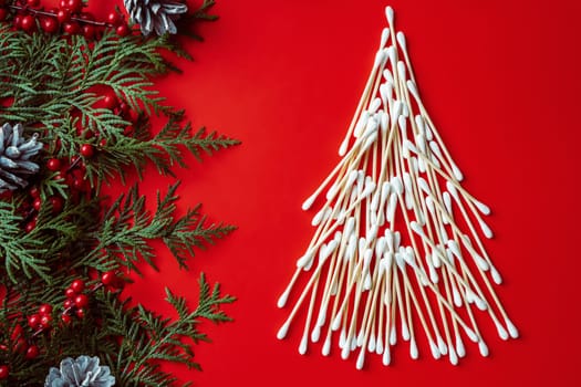 Wooden cotton swabs, laid out in shape of Christmas tree with spruce branches, cones and red berries on red background. Flat lay