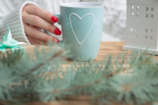 A woman's hand in a white knitted sweater with red nail polish holds a mint mug with a heart, near the branches of a New Year tree and a gift.
