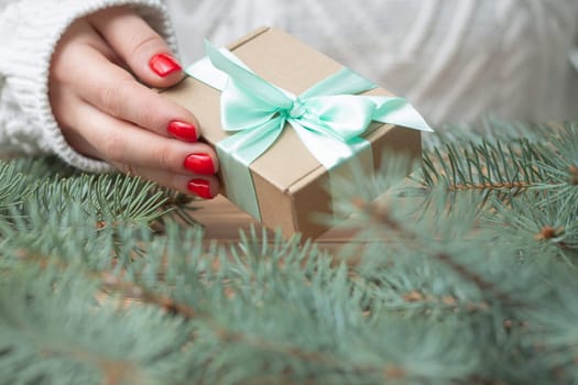 A woman's hand in a white knitted sweater with red nail polish holds a gift with a mint ribbon, near the branches of a Christmas tree.