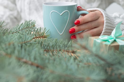 A woman's hand in a white knitted sweater with red nail polish holds a mint mug with a heart, near the branches of a New Year tree and a gift.