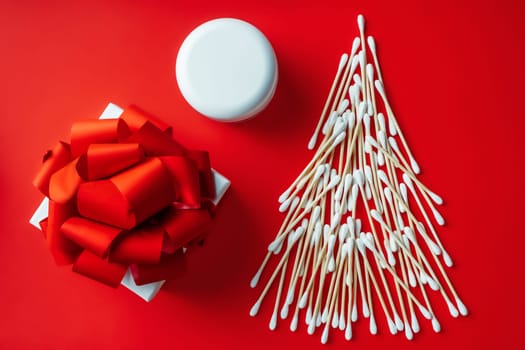 Wooden cotton swabs laid out in shape of Christmas tree with cosmetics cream jar and gift box on red background. Flat lay