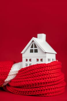 White toy house wrapped in red scarf on red background. Heating or insulating buildings or houses concept. Copy space. Vertical
