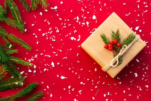 Gift wrapped in craft paper with green branches of a Christmas tree and red berries on a red background. Flat lay. Copy space.