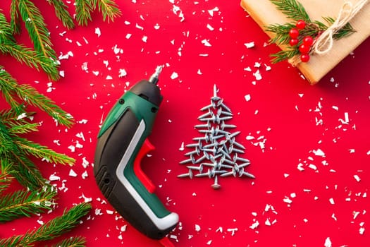 Christmas tree made of self-tapping screws, screwdriver, gift in craft paper with branches of a Christmas tree and red berries on a red background. Flat lay. Copy space.