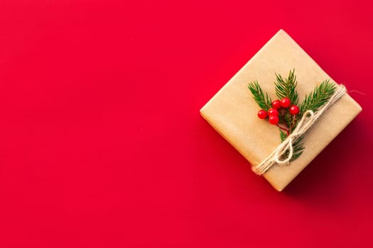 Gift wrapped in craft paper with green branches of a Christmas tree and red berries on a red background. Flat lay. Copy space.