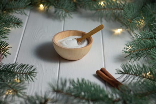 Sugar in a wooden bowl with a spoon and a cinnamon stick next to green branches of a Christmas tree with LED light bulbs garland on a white wooden background. Christmas and New Year baking concept