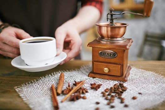 Wooden coffee grinder, coffee beans, star anise, cinnamon sticks on napkin and cup of black coffee in barista hands