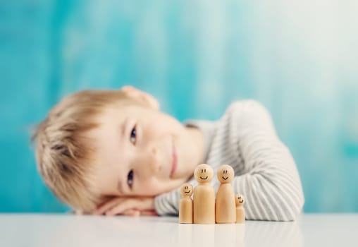 Cute boy sitting at the desk with wooden figures of the family on it. Concept of the love, adoption and relation between peoples.