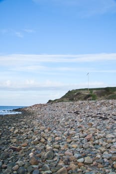 Stone beach by the sea. Flagpole with Danish flag on a hill. Danish coast on a sunny day with clouds in the sky. Landscape shot at the water