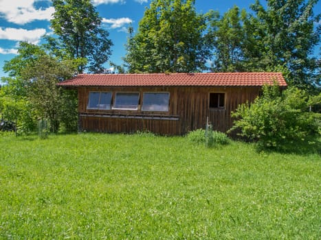 Frontal view of the long side with three glass windows of a beekeeper's house in Bavaria on a green meadow with group of trees in the background under a blue sky with a few white clouds.