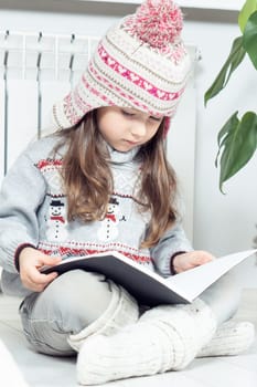 Vertical photo of concentrated, serious, curious cute little girl in woolen hat and warm clothes with snowman reading book with crossed legs next to heating radiator indoors. Study in cold apartment