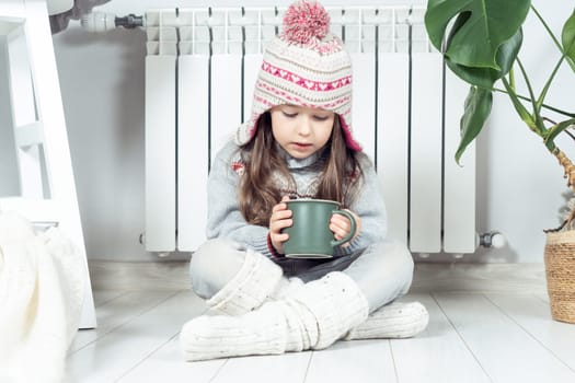 Sad little girl in woolen hat, socks and pullover drink hot tea and sit with crossed legs near heating convector, calorifer. Seasonal sick, virus and flu. Home treatment, healthcare in cold season
