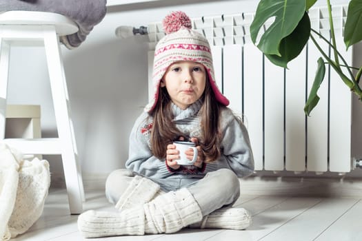 Grimacing little girl in hat, socks and pullover drink sour medicine syrup and sit near heating convector, radiator, next to houseplant. Seasonal sick. Weak throat, immune system in cold winter