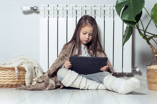 Relaxed little brunette long haired girl using electronic tablet, live streaming online, blogging, vlogging and surfing net, wrapped in blanket, sitting near warm heating radiator and houseplant