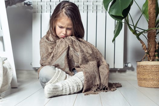 Depressed, cried, serious little girl wrapped in warm plaid, trying warming up, keep warm with woolen clothes, sit on the floor near heating radiator in apartment. Broken convector, suffer from cold