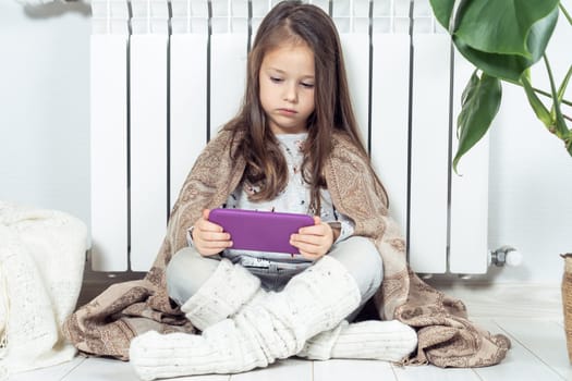 Small little brunette long haired girl using electronic tablet, surfing net and study online, wrapped in blanket, sitting near warm heating radiator. Wireless technology, online school and e learning