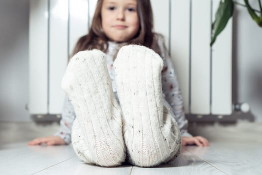 Close up, focus on smiling, calm little girl feet in white woolen socks, showing to camera, sitting near heating radiator at home. Warming up after winter walking, relax. Cozy Christmas holidays
