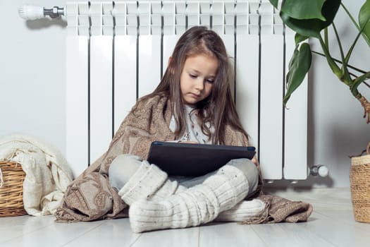 Small cute brunette long haired girl using electronic tablet, watch video, share messages, wrapped in blanket, sitting near warm heating radiator. Wireless technology and digital native generation