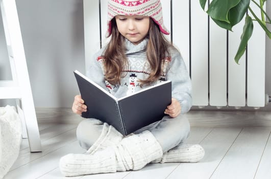 Pensive, serious, cute little girl in woolen hat and warm clothes with snowman reading book or notepad next to heating radiator indoors. Study at home, do homework and learning new on lockdown