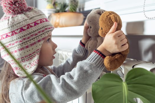 Close up calm, concentrated little girl in knitted hat and sweater warming up with best friends stuffed toys near heating radiator, playing and having fun, care about plushy family. Profile view