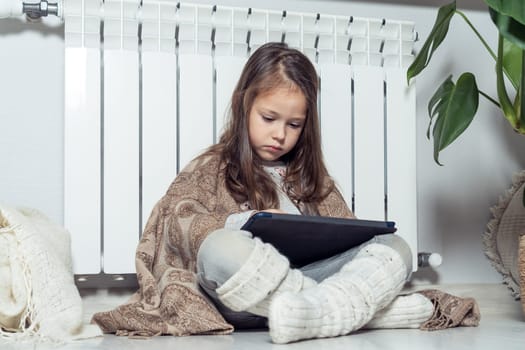 Serious, concentrated little brunette girl use electronic tablet, play games and surfing net in blanket, sit on floor near heat radiator. Wireless technology and modern gadgets on remote location