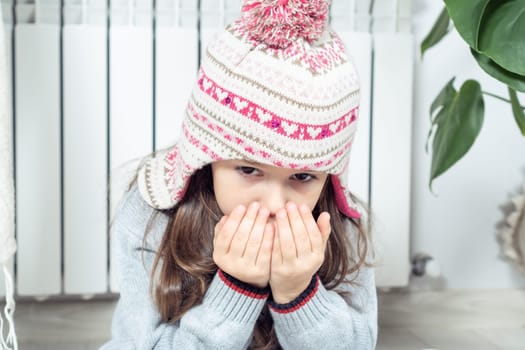 Close up of little brunette girl face breathing on hands to keep warm near cold broken heating radiator, looking at camera indoors. Cold flat in winter season. Poverty and debts, economy and savings