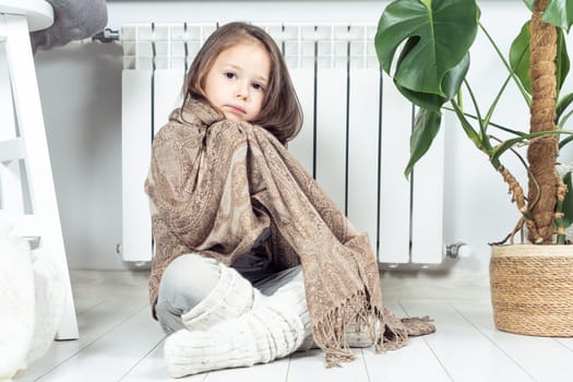 Sad, upset, serious little girl wrapped in warm plaid, keep warm with woolen clothes, look at camera, sit on the floor near heating convector in apartment. Debts and economy, problem central heating
