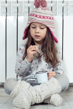Vertical close up little girl in hat, pajamas and socks having lunch, drink hot tea or cocoa and eat sweet cookies, sitting near white heating radiator at home. Cold season and low temperature at home
