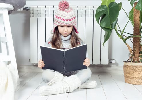 Serious, cute, concentrated little girl in knitted hat and warm clothes reading, exploring book next to heating radiator, near plant in white room. Explore and research of notebook or sketchbook
