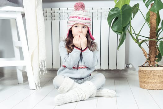Little brunette girl breathing, inhaling on hands to keep warm near cold heating radiator, convector indoors. Cold apartment in winter season. Broken convector, problem with gas, electric radiator