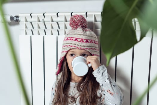 Close up photo of lovely little girl in woolen hat and pajamas drink hot tea and eat cookies, sit near heating radiator at home. Central heating savings, economy. Cold apartment. Plant on foreground