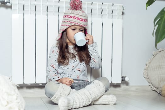 Nice curly little girl in knitted hat and woolen socks eat sweet dessert, drink hot cacao, sitting near white heating radiator in light room. Holidays in winter, hygge and comfort in cold season