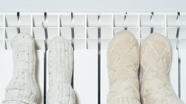 Close up photo of cute, cozy child and adult raising legs in wool, knitted socks warm themselves near heat radiator. Central heating, gas consumption in cold winter season. Christmas evening at home