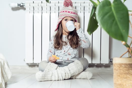 Cute little girl in knitted hat and socks have lunch, drink hot tea or cacao and eat sweet cookies, sitting near white heating radiator at home. Holidays in winter, cold season and low temperature