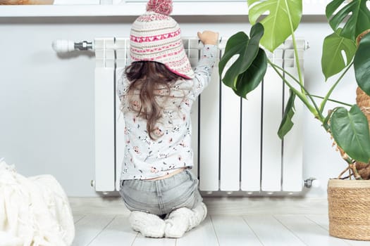 Rearview of frozen little curly girl in warm knitted hat sitting on knees and warm hands on heat radiator. Safety and protection at home. Gas and heating saving. Climate change and low temperature