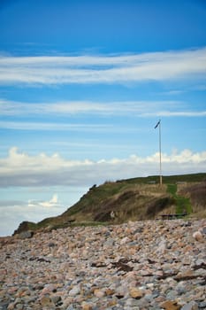 Stone beach by the sea. Flagpole with Danish flag on a hill. Danish coast on a sunny day with clouds in the sky. Landscape shot at the water