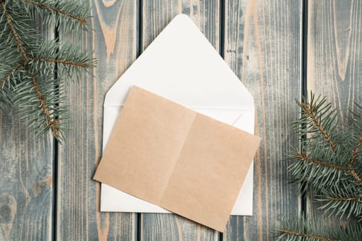 White open envelope on gray wooden background with blank kraft paper greeting card with branches of Christmas tree. New year envelope. Copy space. Top view