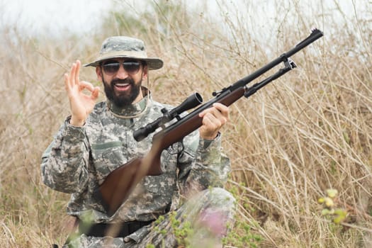 The smiling hunter in military uniform with rifle is showing OK with his hand outdoors.