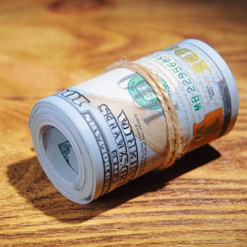 A roll of dollars on a wooden textured table in the dark illuminated by a ray of light