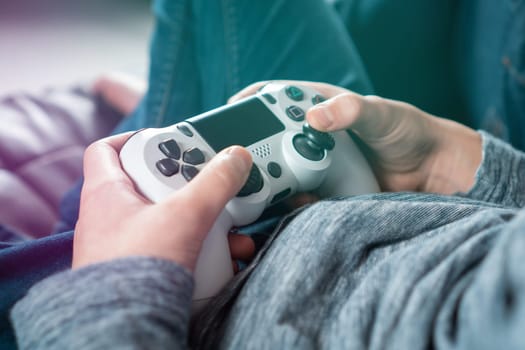 Cropped photo of teenage boy sitting at home, holding white gaming controller joystick gamepad, playing videogames on console station. Hobby, free time, gaming, entertainment, leisure. Soft focus.