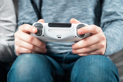 Unrecognizable teenage boy child sitting at home, holding white gaming controller joystick gamepad, playing videogames on game console. Hobby, free time, gaming, interactive entertainment, leisure.