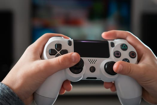 Unrecognizable teenage boy child sitting at home, holding white gaming controller joystick gamepad, playing videogames on console station. Hobby, free time, gaming, entertainment, leisure. Soft focus.
