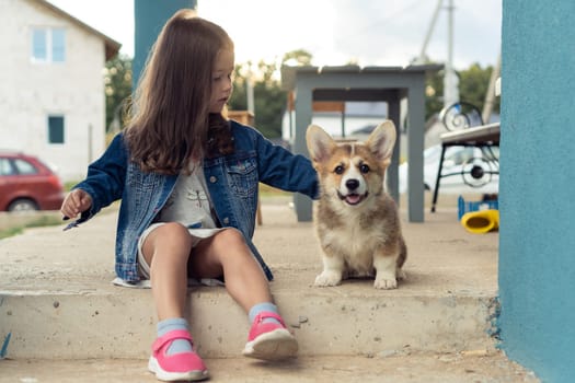 Portrait of wonderful little girl with long dark hair wearing denim jacket, caressing, stroking fur of happy welsh pembroke corgi puppy pet, sitting on concrete stairs in yard. Pet care, animal life.