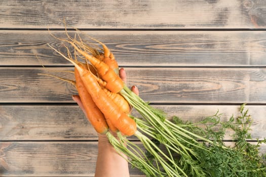 Unrecognizable cropped man hand holding orange tasty juicy carrot with green stem and leaves on wooden background. Ecological lifestyle, carotene vitamins in organic food. Top view, copy space